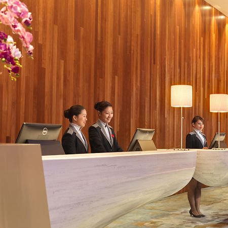 马来西亚大红花（丽昇精选酒店） 波德申 外观 照片 The photo showcases a hotel reception area. Three women in professional attire are standing behind a sleek, modern reception desk. The desk has a unique design, and there are computer monitors positioned on it. The backdrop features a warm wooden wal