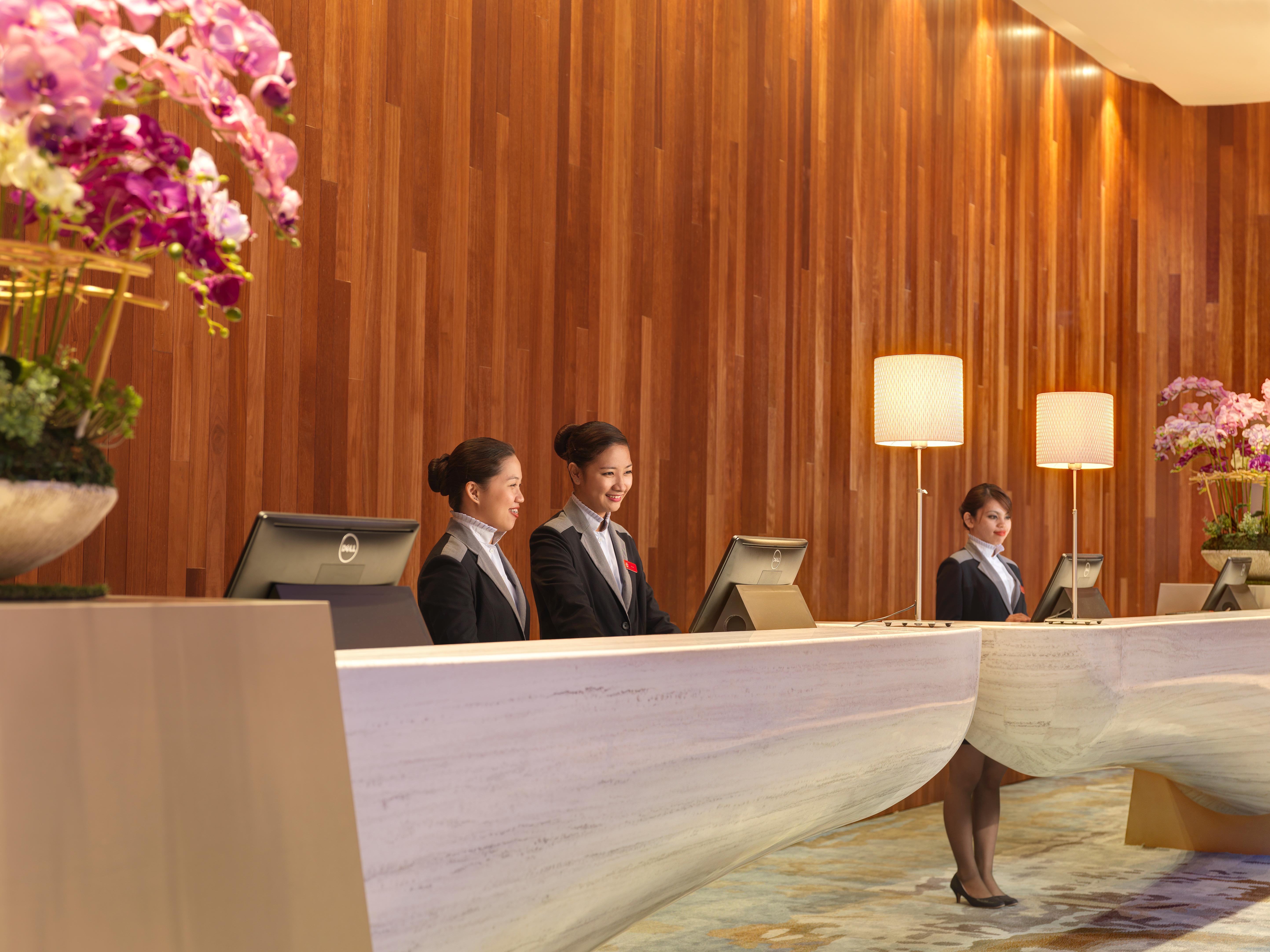 马来西亚大红花（丽昇精选酒店） 波德申 外观 照片 The photo showcases a hotel reception area. Three women in professional attire are standing behind a sleek, modern reception desk. The desk has a unique design, and there are computer monitors positioned on it. The backdrop features a warm wooden wal